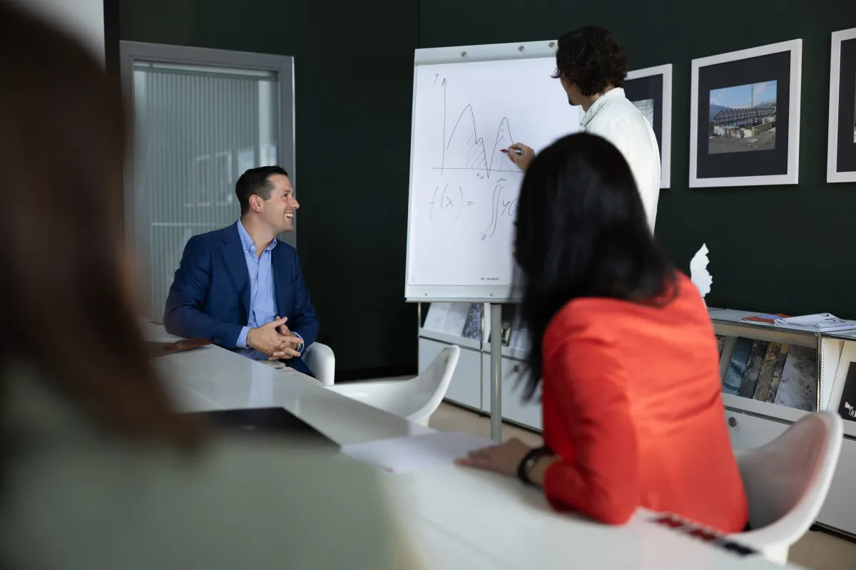 Teilnehmende bei einer Präsentation im Seminarraum, Fokus auf Vortragendem und Flipchart mit Funktionsgleichung.