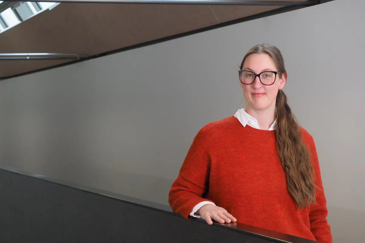 Eine Person mit Brille und langem Haar steht in einem modernen Gebäude und trägt einen roten Pullover. Sie lächelt leicht und lehnt sich an ein Geländer.