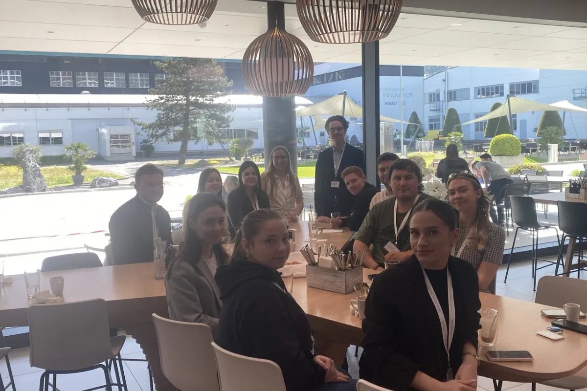 Studierende der Universität Liechtenstein sitzen in einer modernen Cafeteria bei Ivoclar in Schaan. Sie sind um einen Tisch versammelt und blicken in die Kamera. Im Hintergrund sind große Fenster mit Blick auf den Außenbereich zu sehen.