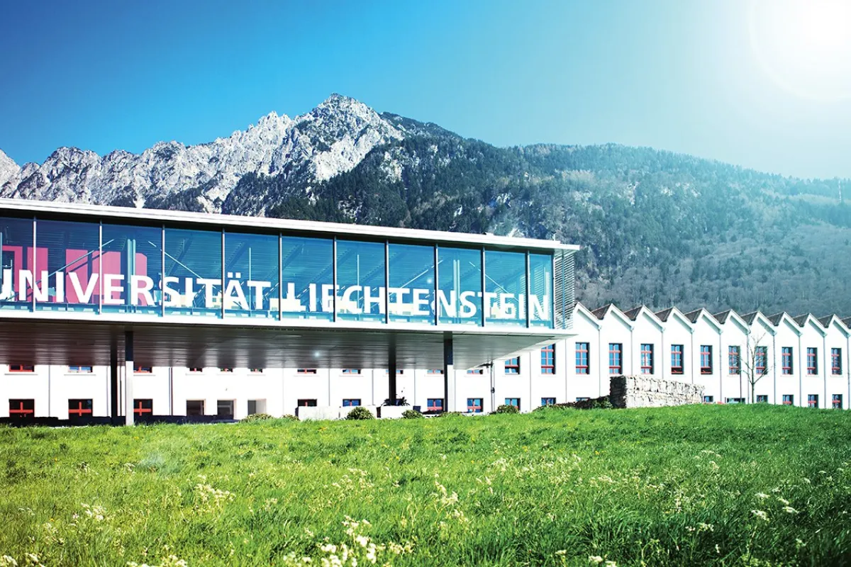 Außenansicht der Universität Liechtenstein mit modernem Glasgebäude und Bergkulisse im Hintergrund, umgeben von grüner Wiese.