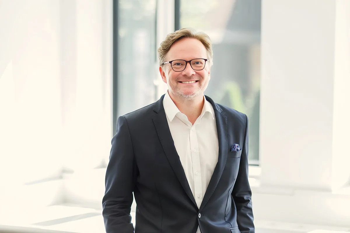 Ein lächelnder Mann in einem Anzug steht vor einem hellen Fenster.