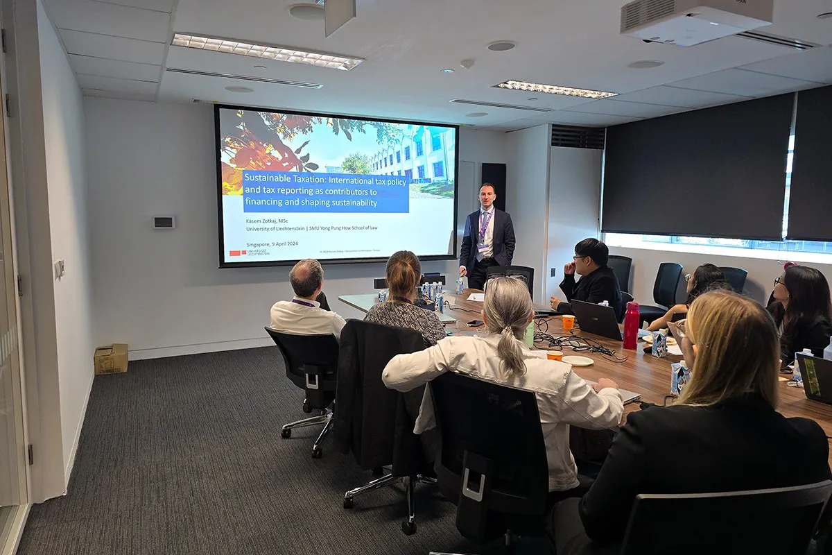 Ein Vortragender steht vor einer Präsentation mit dem Titel "Sustainable Taxation" in einem Konferenzraum. Mehrere Personen sitzen an einem Tisch und hören zu. Die Präsentation behandelt internationale Steuerpolitik und Nachhaltigkeit.
