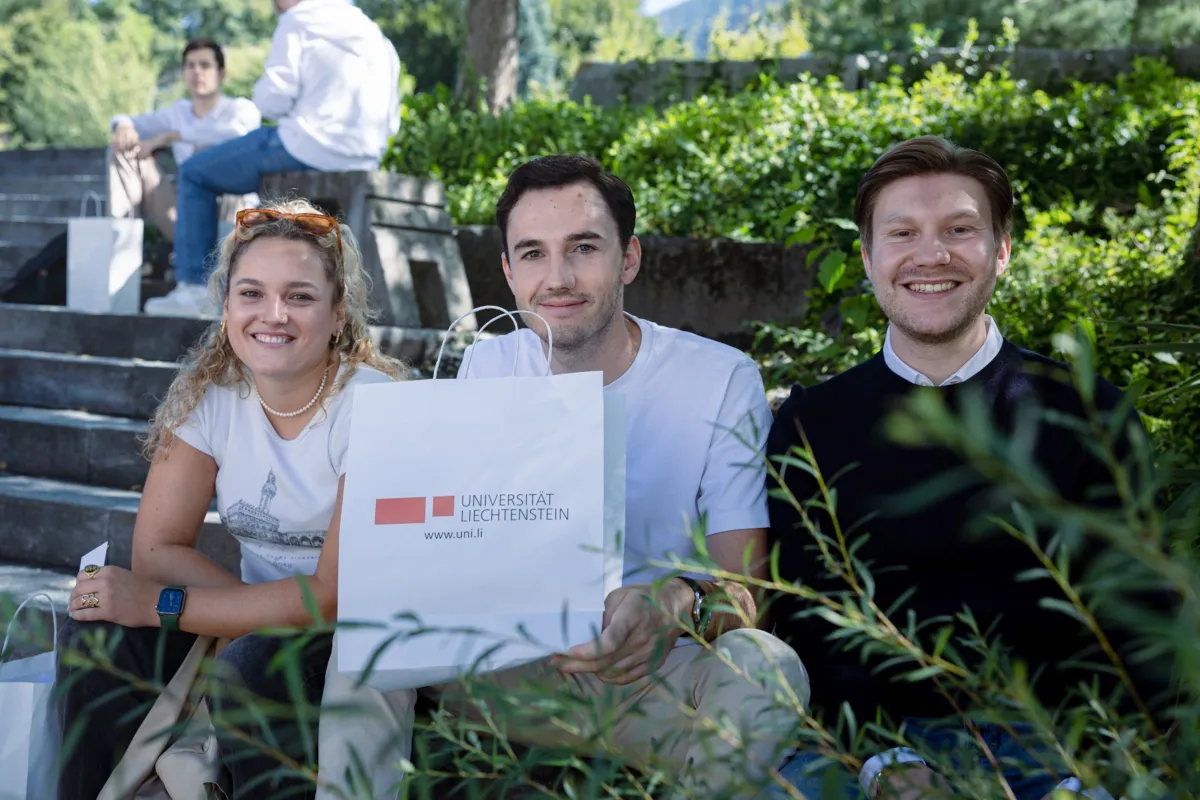 Drei neue Studierende sitzen auf dem Vorplatz des Campus Universität Liechtenstein und haben eine Tasche mit dem Logo der Universität in der Hand