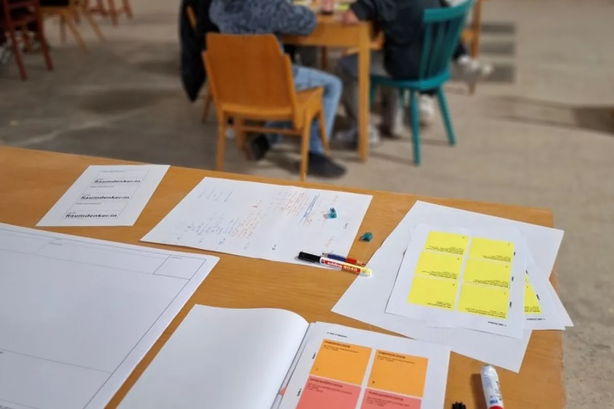 Blick auf einen Schreibtisch mit bunten Notizzetteln und Menschen, die an einem Tisch sitzen im Hintergrund