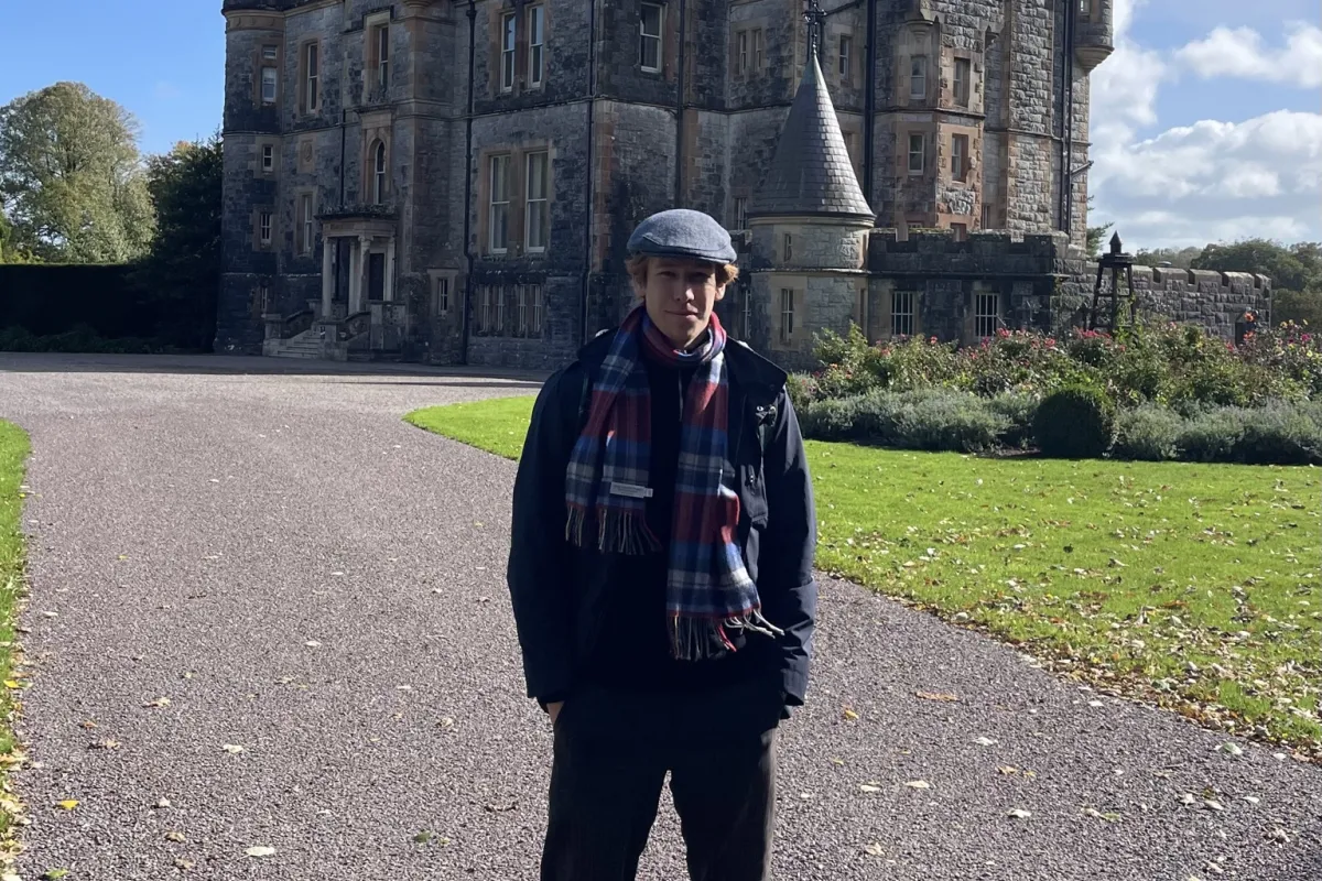 Person mit Mütze und Schal steht auf einer Auffahrt vor einem historischen Schloss aus Stein unter blauem Himmel.