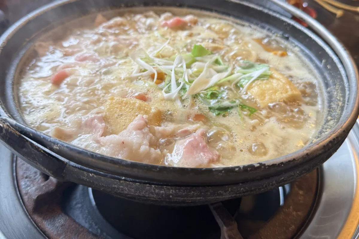 Dampfender Hot Pot mit Fleisch, Tofu und Gemüse in heller Brühe auf einem Tisch-Gasbrenner.