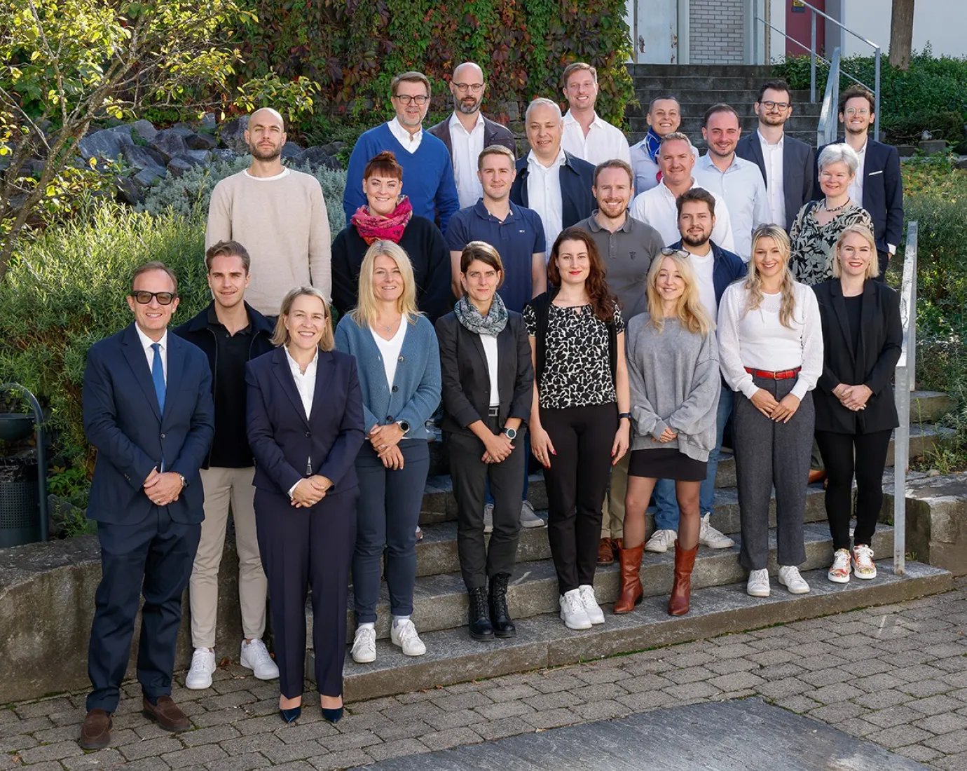 Eine Gruppe von Studierenden und Dozenten des LL.M.-Executive-Masterstudiengangs im Gesellschafts-, Stiftungs- und Trustrecht der Universität Liechtenstein posiert auf einer Treppe im Freien. Sie sind Teil des I&F Family Wealth Preservation Awards. Im Hintergrund sind Pflanzen und eine Gebäudefassade zu sehen.