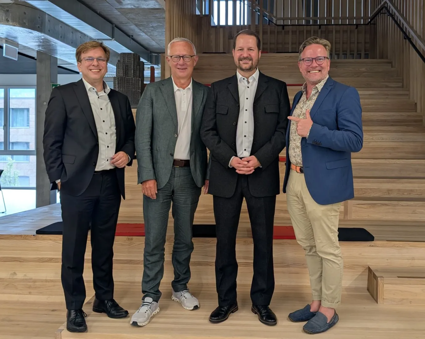 Vier Männer in formeller Kleidung stehen lächelnd auf einer Holztreppe in einem modernen Gebäude.