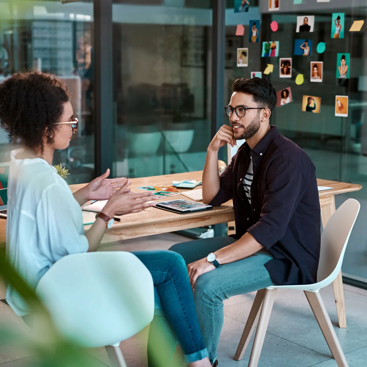 Eine Frau und ein Mann sitzen sich in einem modernen Büro gegenüber. Die Frau spricht gestikulierend, während der Mann aufmerksam zuhört. Auf dem Tisch liegen ein Laptop und Büromaterialien. Im Hintergrund ist eine Glaswand mit bunten Haftnotizen und Fotos zu sehen.