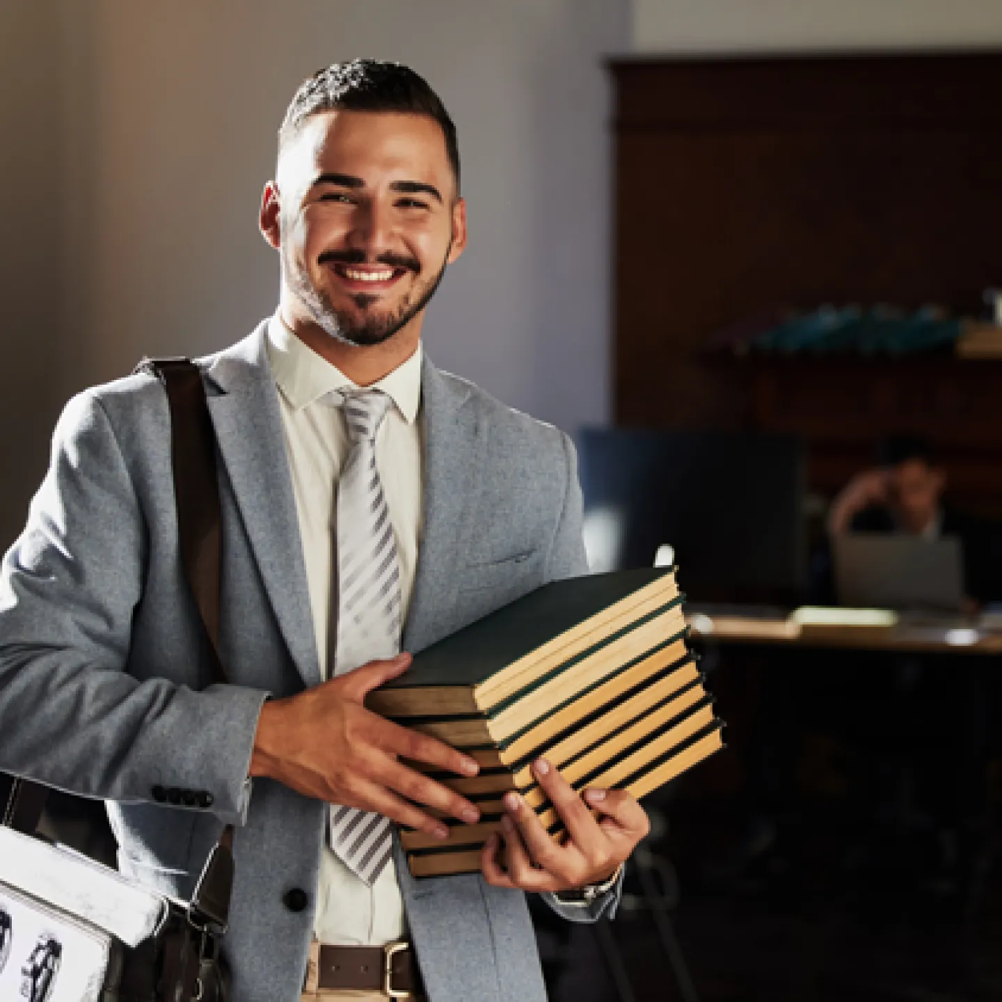 Ein junger, selbstbewusster Mann in einem Business-Anzug, der mehrere Bücher trägt, lächelt freundlich in die Kamera. Der Hintergrund zeigt eine ruhige Büroumgebung, die eine produktive und professionelle Atmosphäre vermittelt.