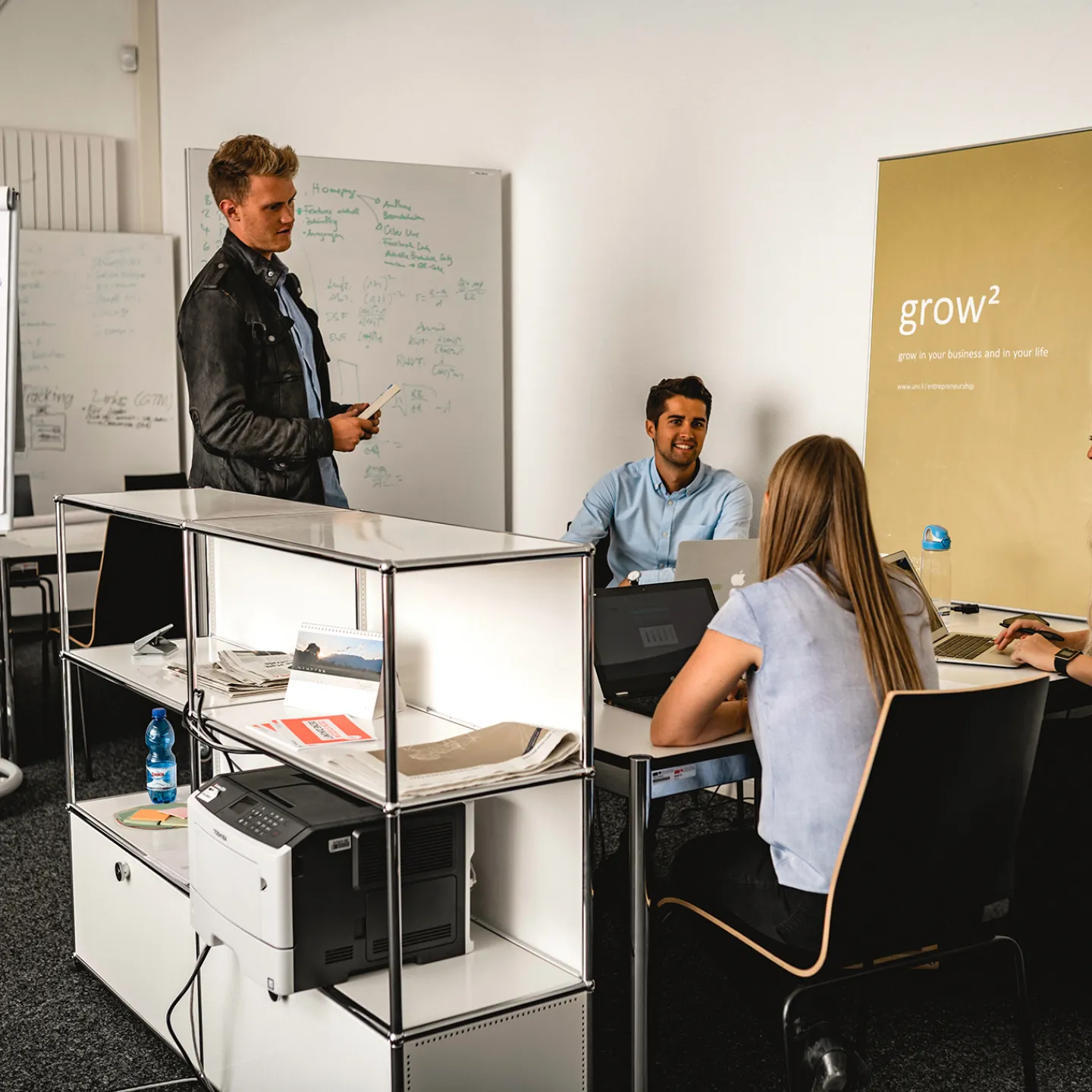 Vier Studierende arbeiten im Team an einem Projekttisch, mit Whiteboard und Flipchart im Hintergrund