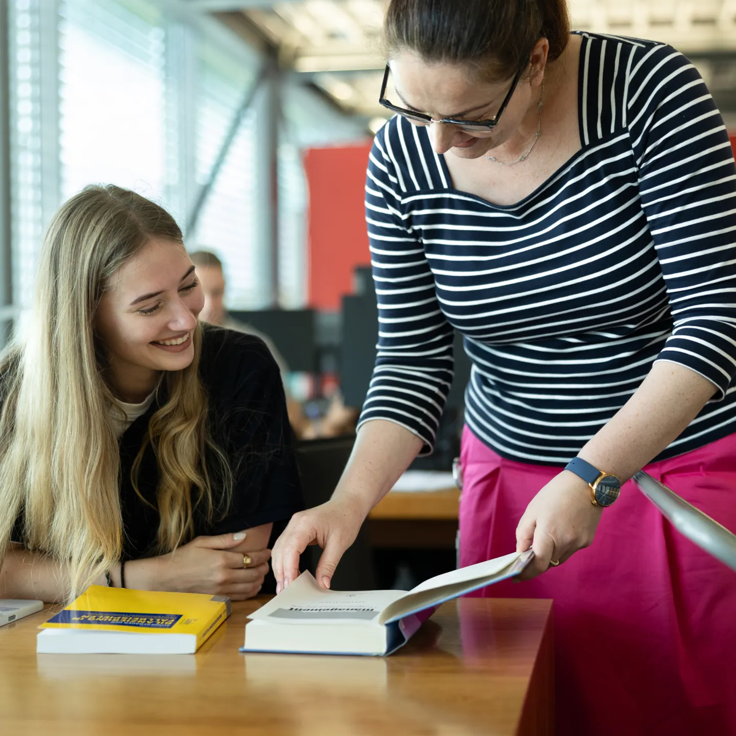 Eine Bibliotheksmitarbeiterin zeigt einer lächelnden Studentin Inhalte aus einem geöffneten Buch.