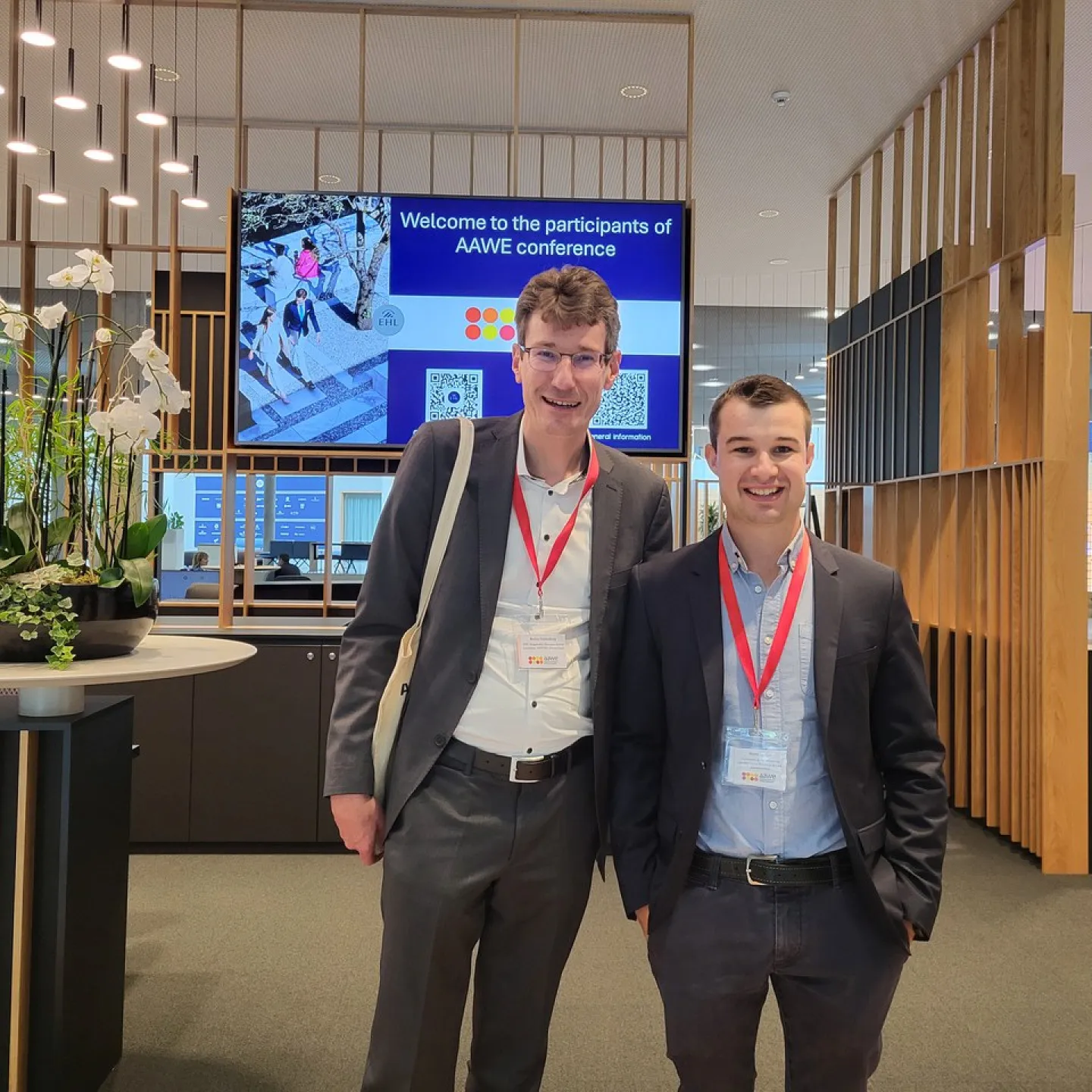 Zwei Männer in Anzügen mit roten Namensschildern stehen lächelnd in einem modernen Konferenzraum. Im Hintergrund ist ein Bildschirm mit der Aufschrift „Welcome to the participants of AAWE conference“ zu sehen. Links steht eine dekorative Pflanze auf einem Tisch.