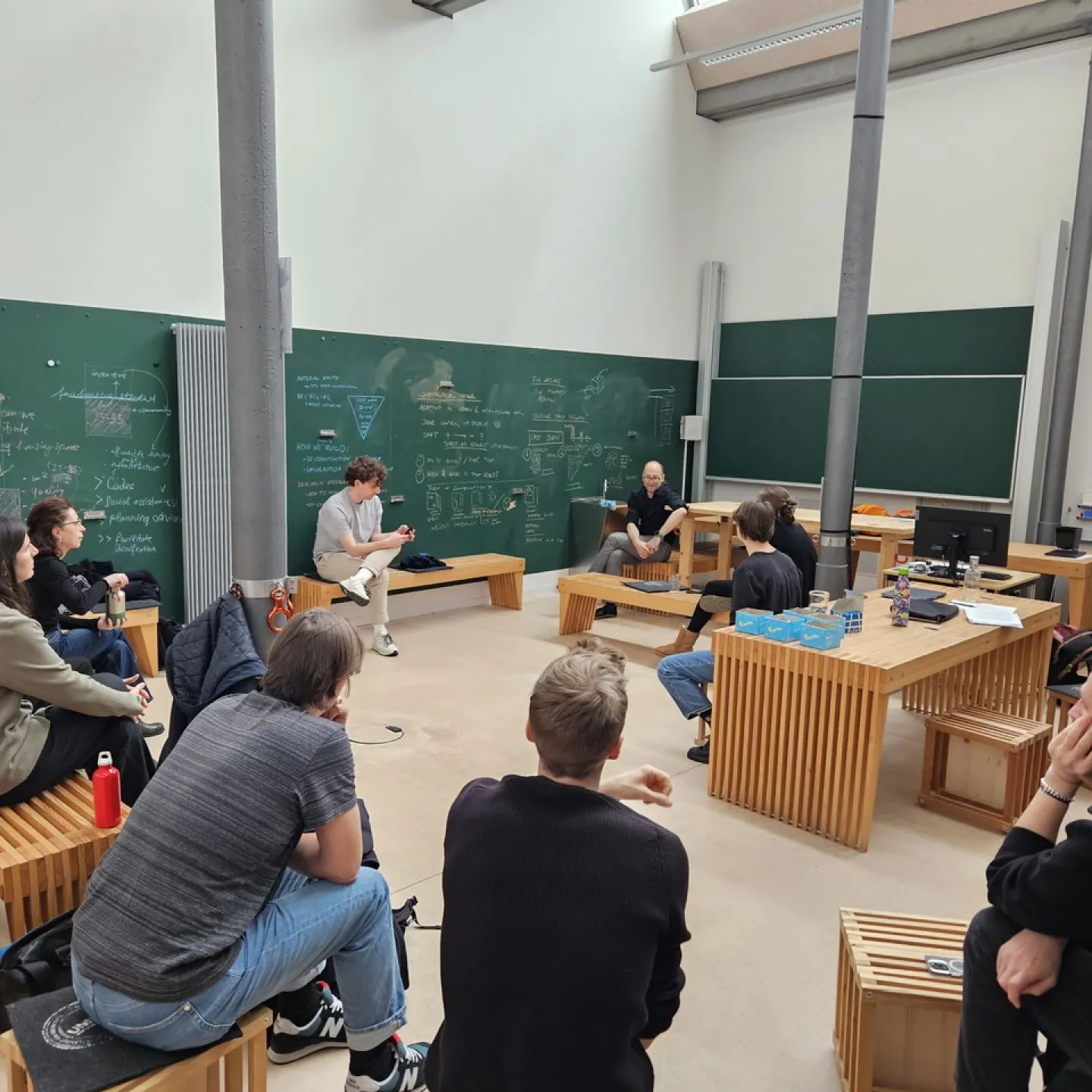 Eine Gruppe von Studierenden sitzt in einem modernen Seminarraum der Universität Liechtenstein, umgeben von Tafeln mit Notizen und Skizzen. Sie diskutieren angeregt, während ein Dozent vorne sitzt und zuhört.