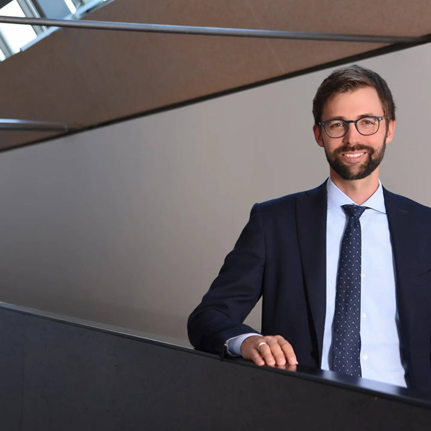 Ein Mann im Anzug steht lächelnd in einem modernen Gebäude mit schrägen Fenstern.