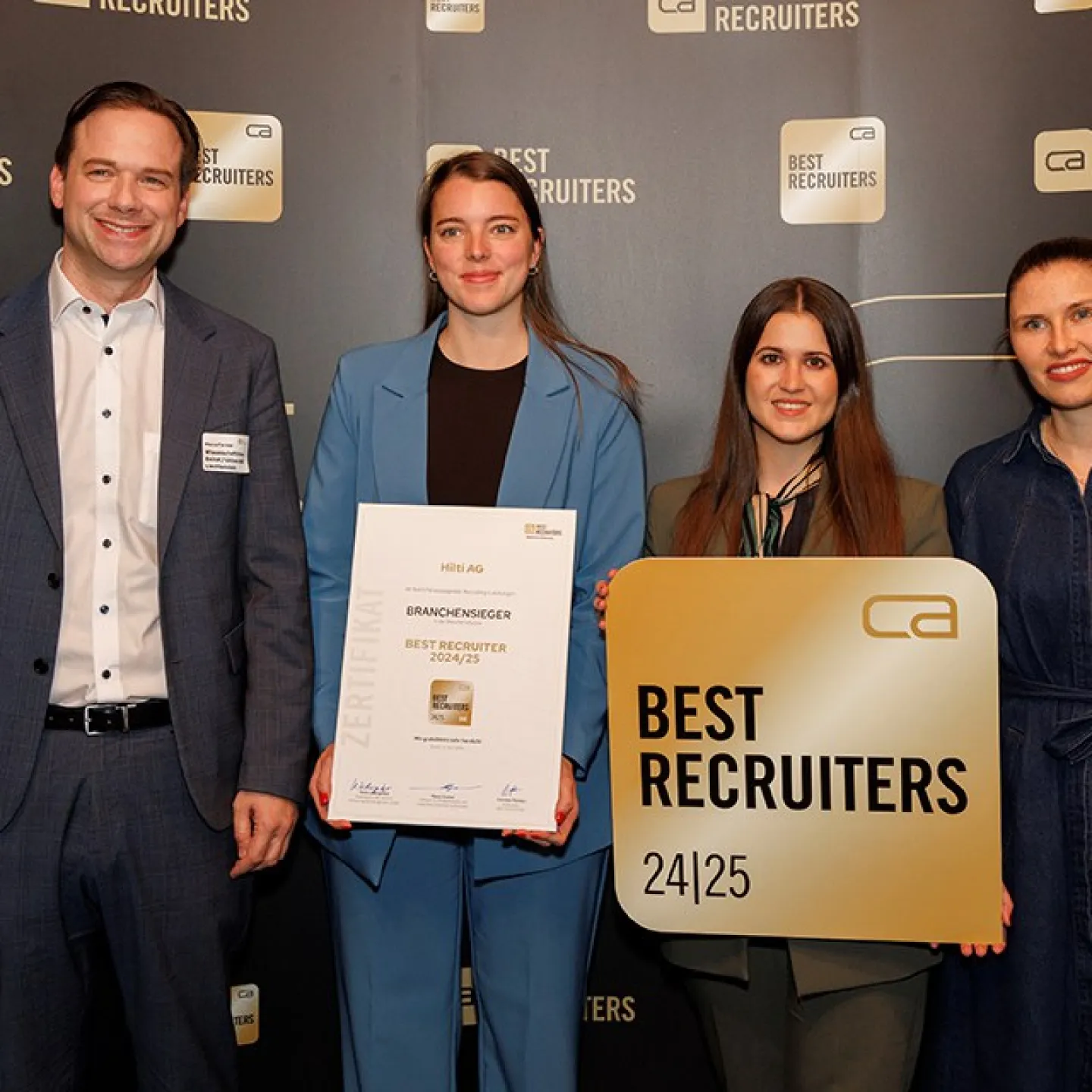 Vier Personen stehen vor einem Hintergrund mit dem Logo "Best Recruiters". Eine Person hält ein Zertifikat mit der Aufschrift "Branchen­sieger Best Recruiter 2024/25". Eine andere Person hält ein Schild mit dem gleichen Logo. Alle lächeln in die Kamera.