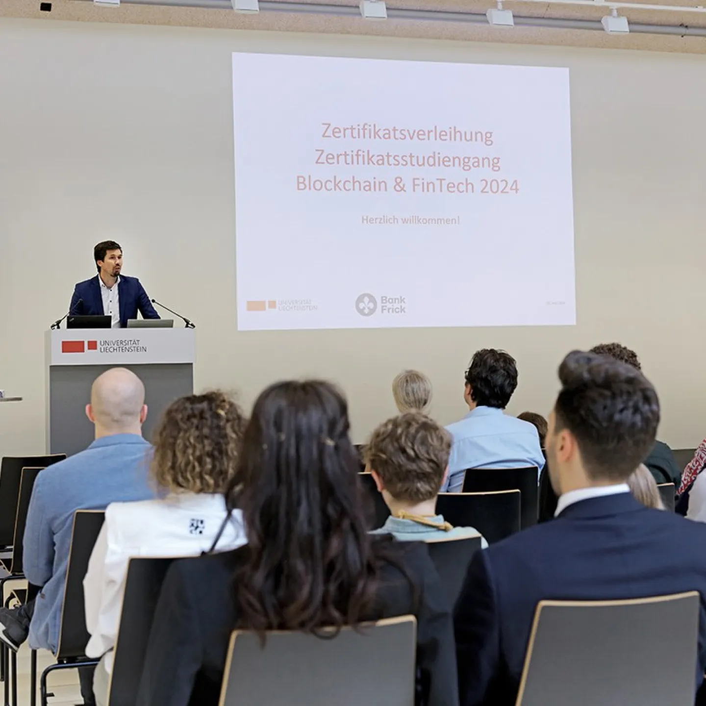 Ein Redner steht an einem Podium vor einer Leinwand mit der Aufschrift "Zertifikatsverleihung Zertifikatsstudiengang Blockchain & FinTech 2024" bei der Universität Liechtenstein. Im Publikum sitzen mehrere Personen. Links und rechts sind Banner von Bank Frick zu sehen.