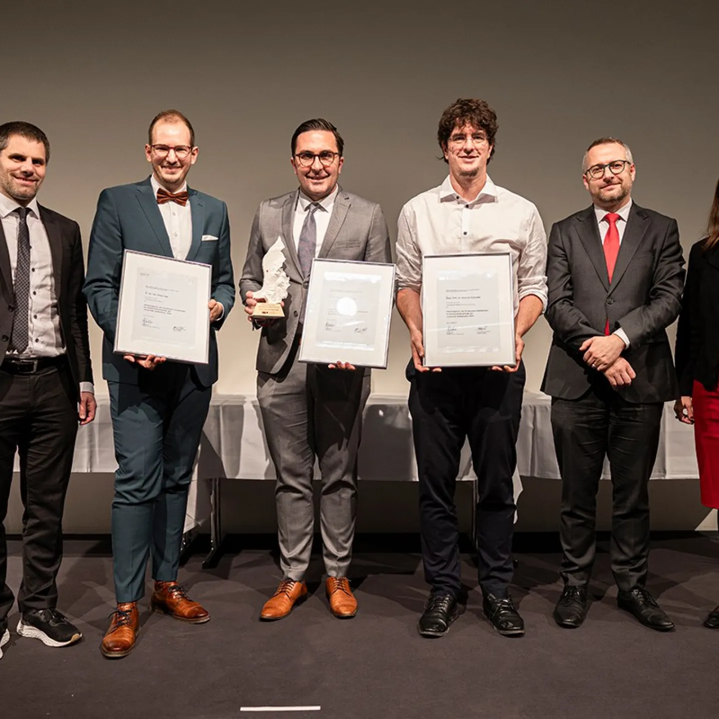 Eine Gruppe von sechs Personen steht nebeneinander auf einer Bühne. Drei Personen in der Mitte halten Auszeichnungen und Urkunden. Alle sind formell gekleidet und lächeln in die Kamera. Im Hintergrund sind weiße Tischdecken sichtbar.