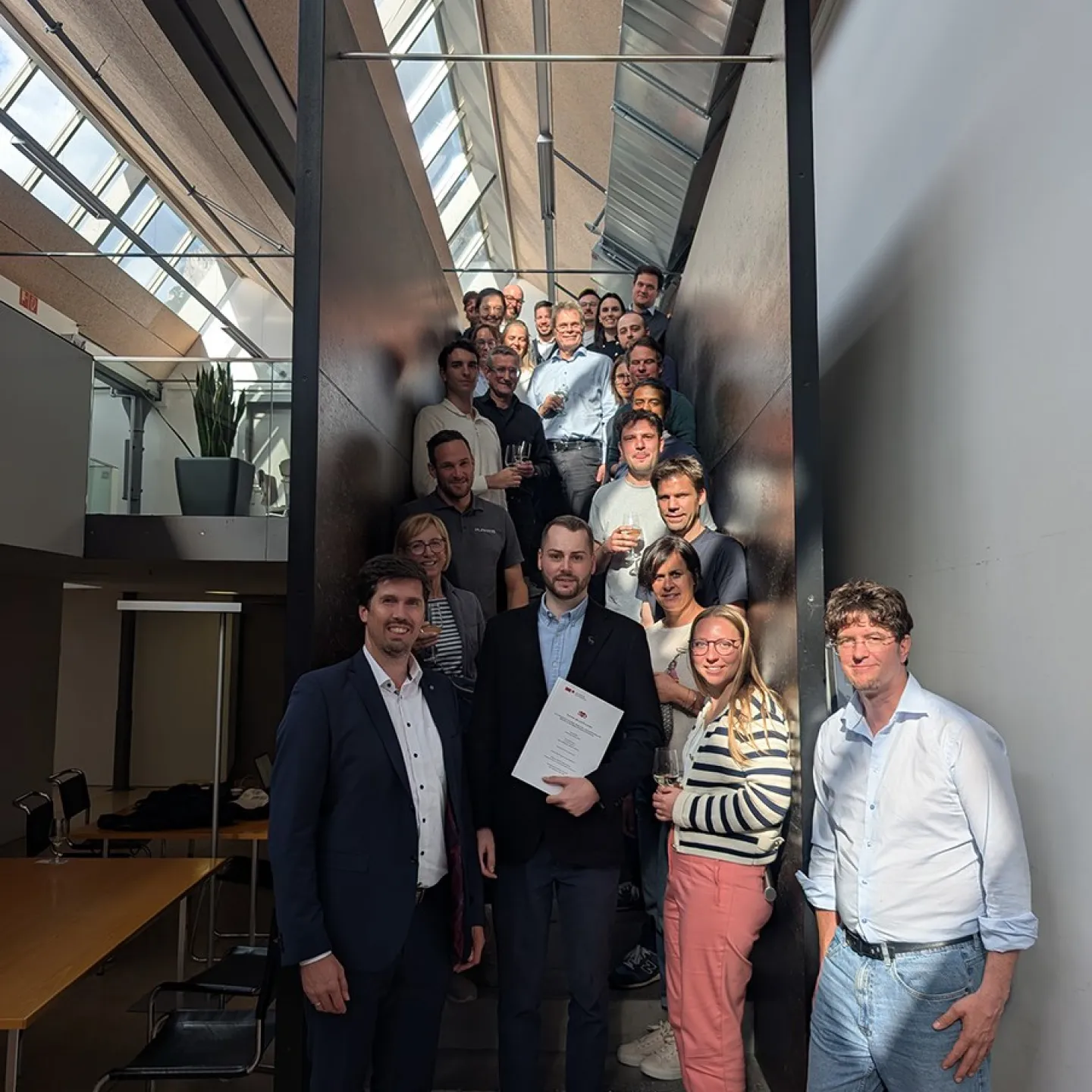 Eine Gruppe von Menschen steht auf einer Treppe in einem modernen Gebäude. Im Vordergrund hält eine Person ein Dokument in der Hand, umgeben von lächelnden Kollegen. Die Szene ist gut beleuchtet durch ein Oberlicht.