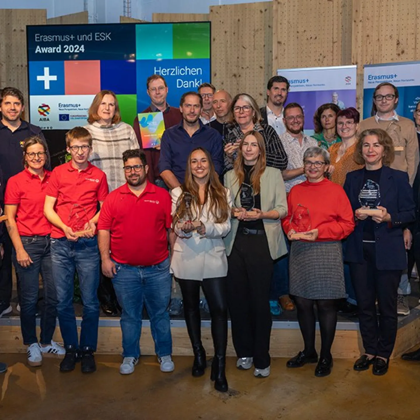 Eine Gruppe von Menschen steht auf einer Bühne und hält Trophäen. Im Hintergrund sind Banner mit Erasmus+ Logos und ein Bildschirm mit der Aufschrift "Erasmus+ und ESK Award 2024" zu sehen. Die Personen lächeln und posieren für ein Gruppenfoto.