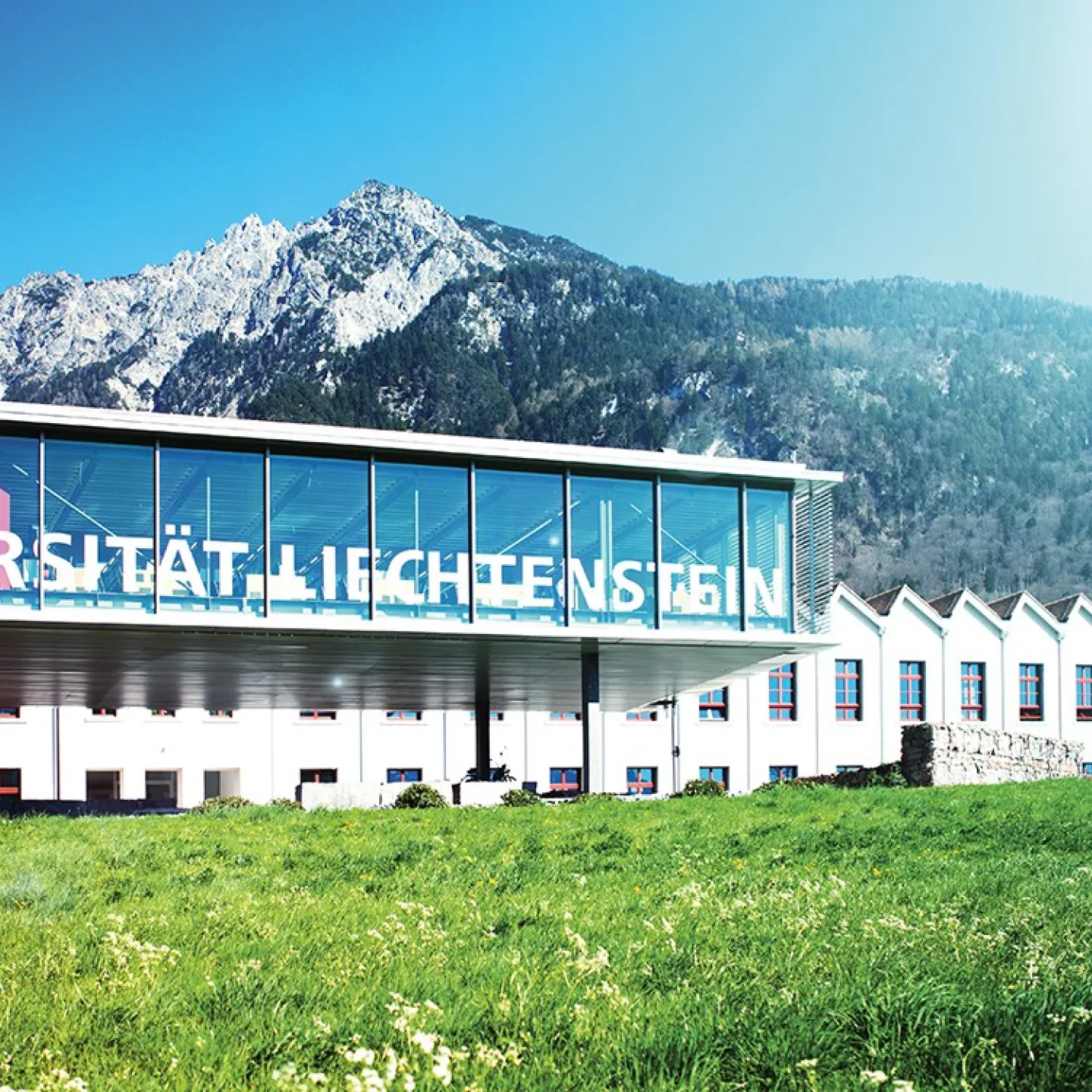 Außenansicht der Universität Liechtenstein mit modernem Glasgebäude vor malerischer Bergkulisse, umgeben von grüner Wiese.