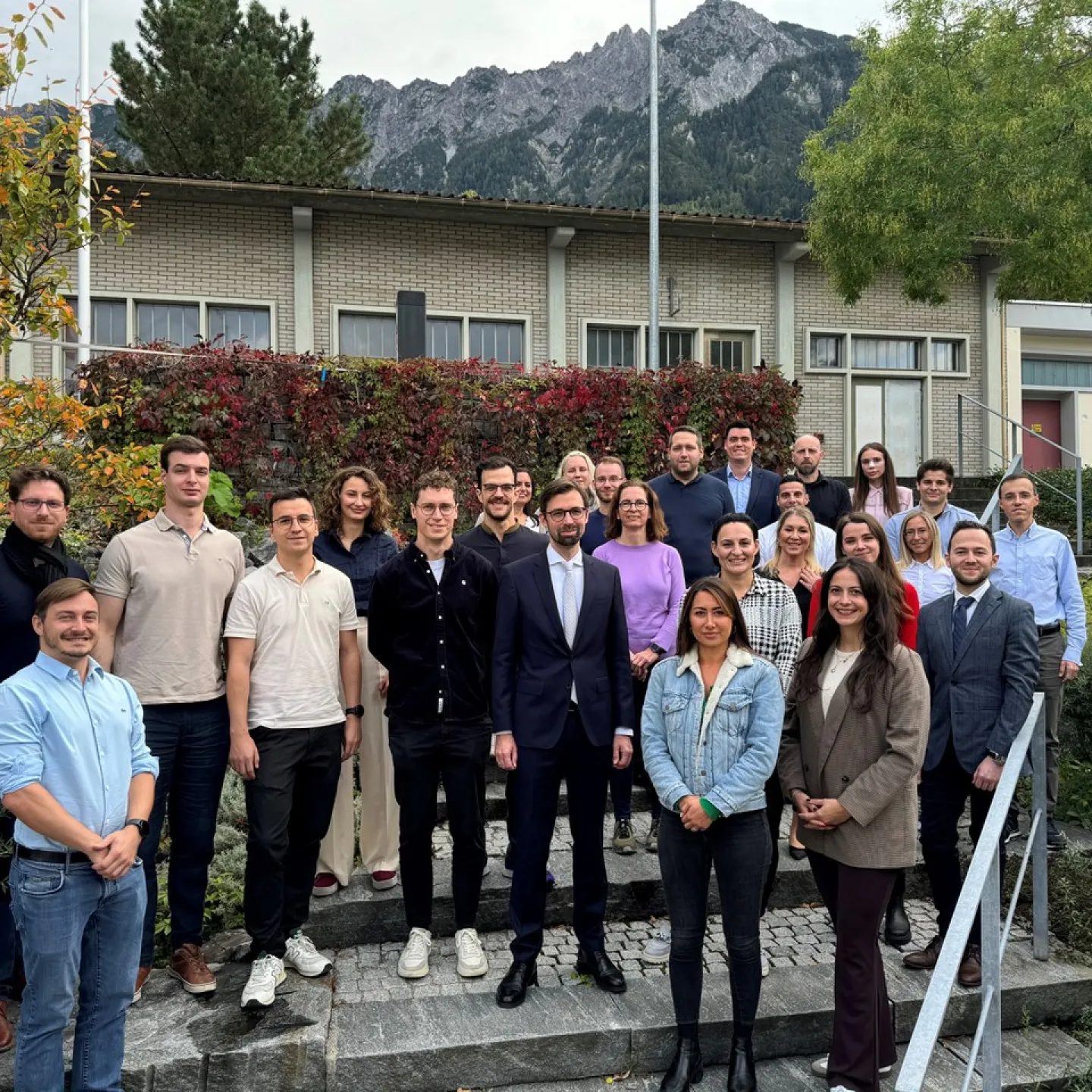 Gruppe von 22 Studierenden und einem Dozenten des Executive Master of Laws (LL.M.) im Bank- und Finanzmarktrecht der Universität Liechtenstein, posierend vor einem Gebäude mit herbstlicher Vegetation und Bergkulisse.