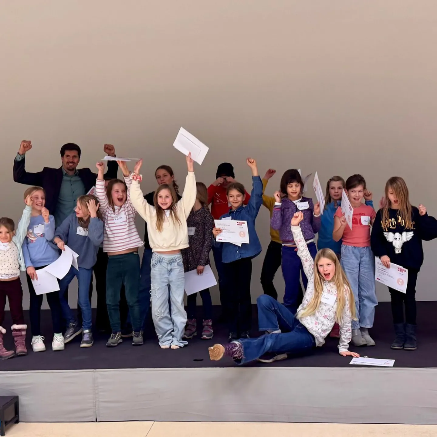Eine Gruppe von Kindern und einem Erwachsenen steht auf einer Bühne und hält stolz Zertifikate hoch. Die Kinder wirken fröhlich und begeistert. Sie nehmen an einem Fundraising-Workshop an der Universität Liechtenstein teil.