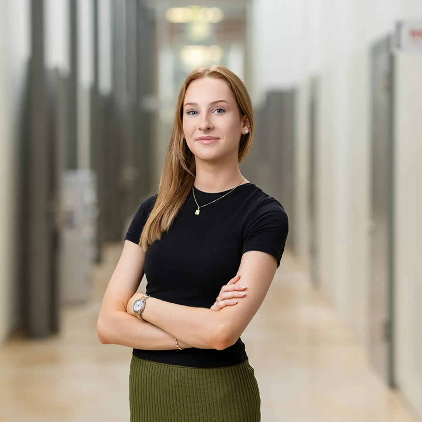 Eine junge Frau steht selbstbewusst mit verschränkten Armen in einem modernen Flur. Sie trägt ein schwarzes Oberteil und einen grünen Rock.