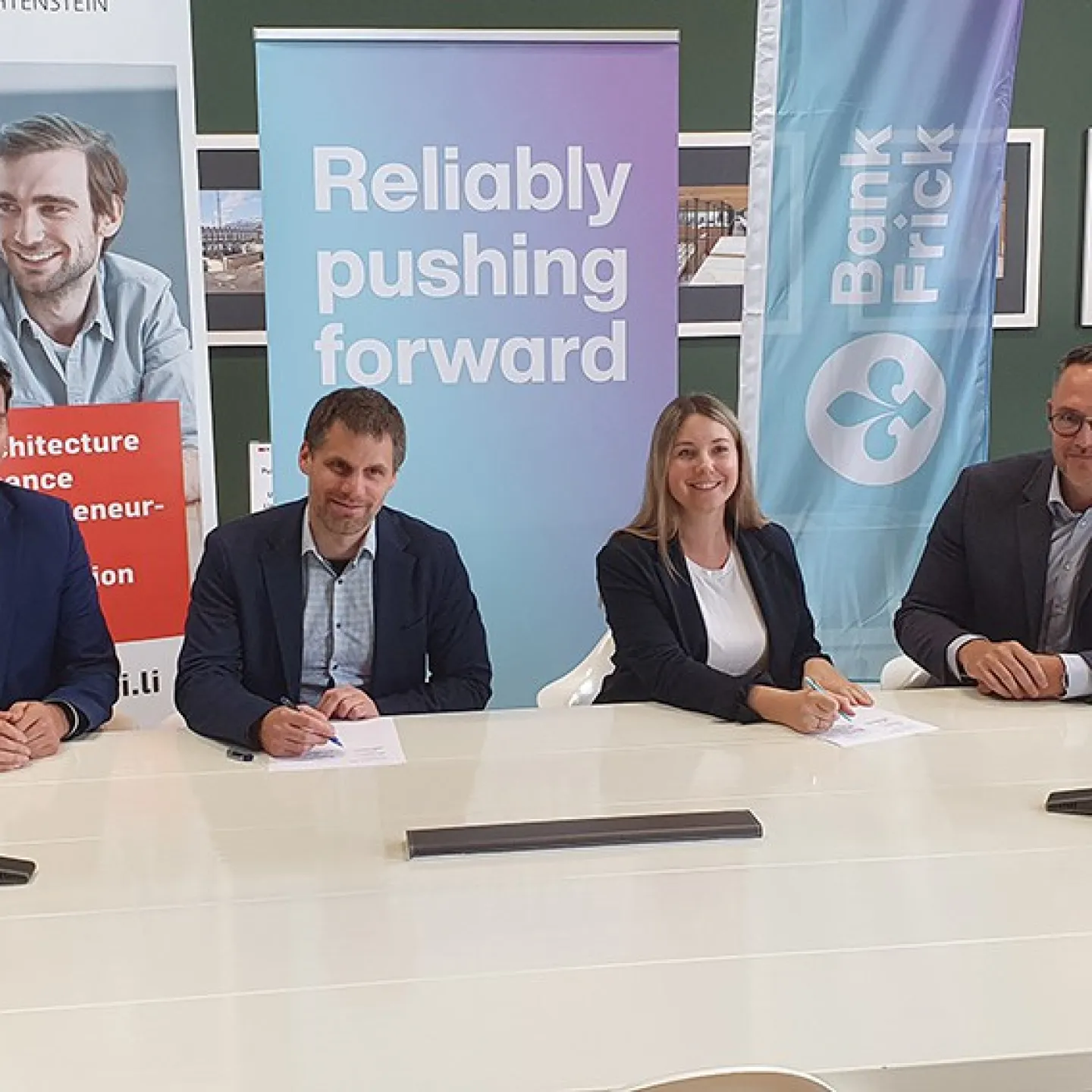 Vier Personen sitzen an einem Konferenztisch und unterzeichnen Dokumente. Im Hintergrund sind Banner der Universität Liechtenstein und Bank Frick zu sehen. Die Szene symbolisiert die Verlängerung der Kooperation zwischen der Universität und der Bank.