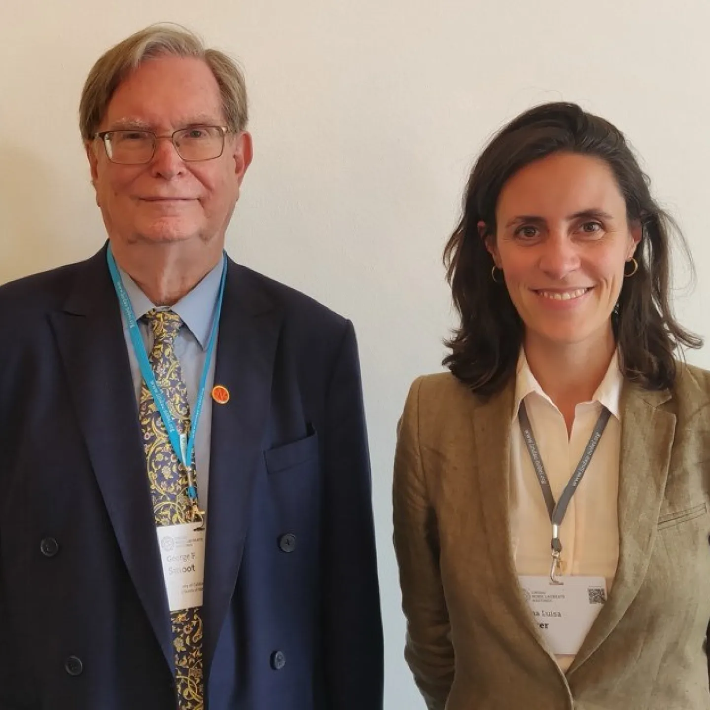 Zwei Personen stehen nebeneinander und lächeln in die Kamera. Beide tragen Namensschilder und sind formell gekleidet.