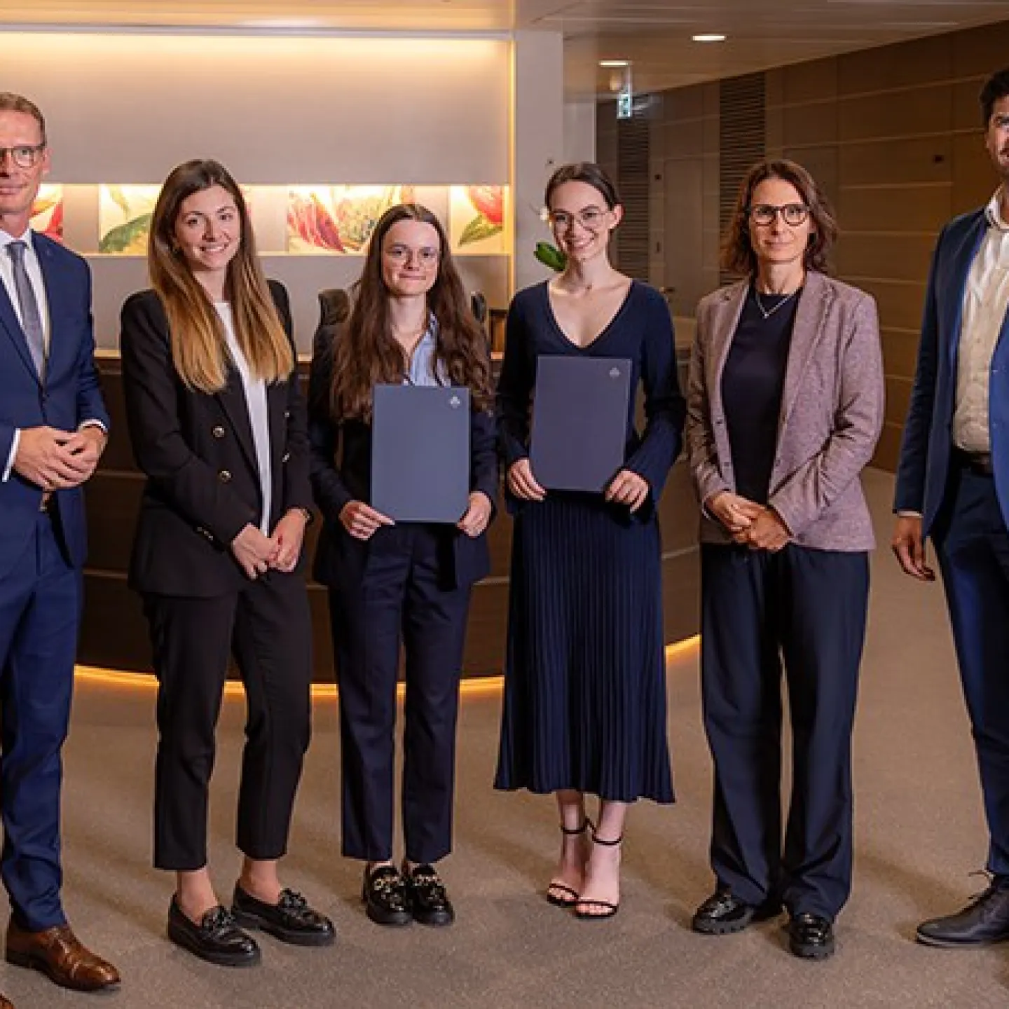 Eine Gruppe von sechs Personen steht in einem modernen Bürogebäude. Zwei Frauen in der Mitte halten Zertifikate und lächeln. Sie werden von vier weiteren Personen flankiert, die ebenfalls lächeln. Die Szene vermittelt eine feierliche Atmosphäre, möglicherweise im Rahmen einer Stipendienverleihung an der Universität Liechtenstein.