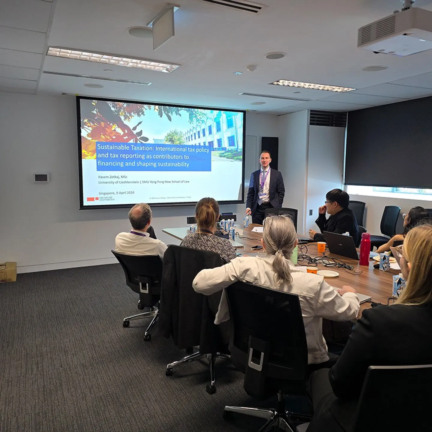 Ein Vortragender steht vor einer Präsentation mit dem Titel "Sustainable Taxation" in einem Konferenzraum. Mehrere Personen sitzen an einem Tisch und hören zu. Die Präsentation behandelt internationale Steuerpolitik und Nachhaltigkeit.