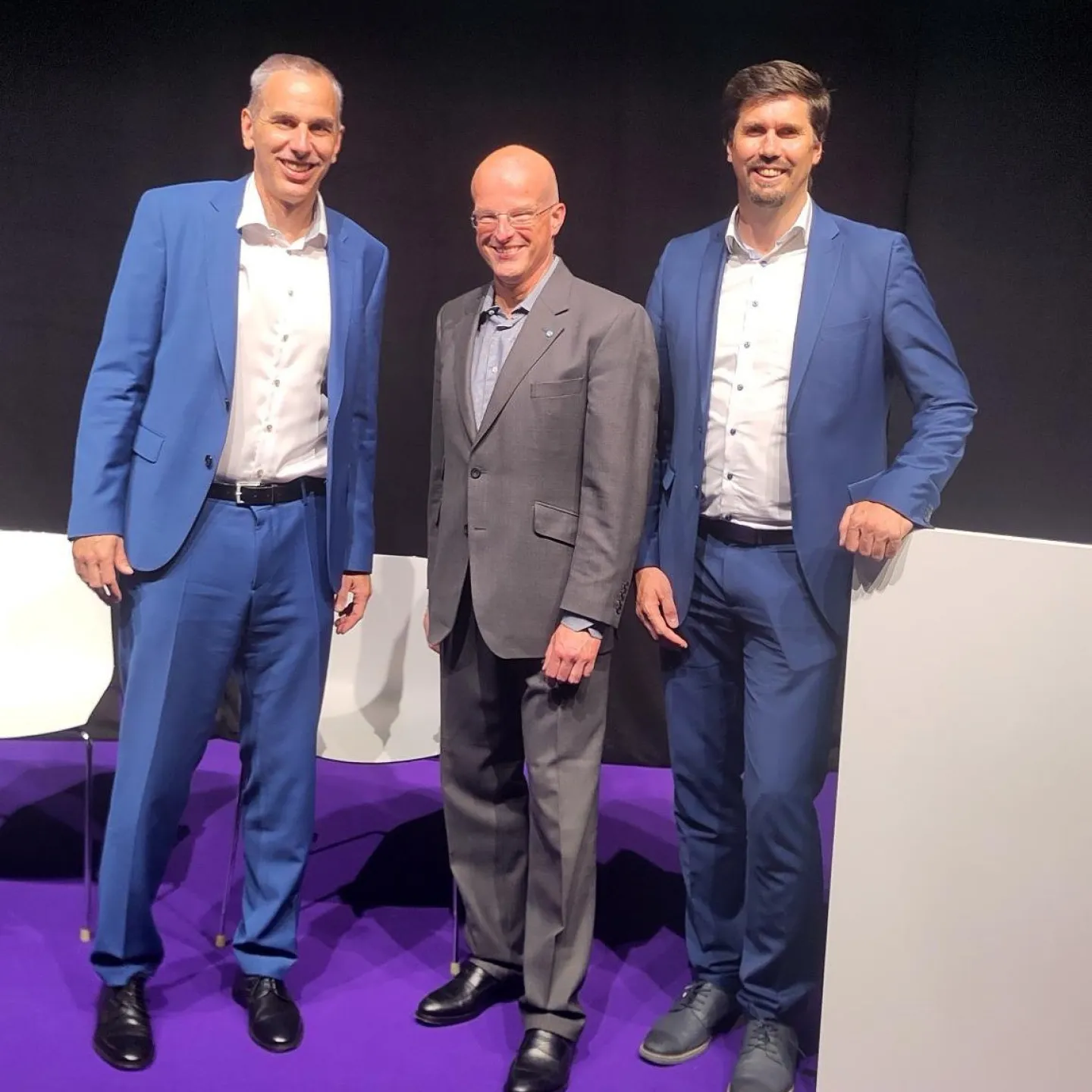 Drei Männer in Anzügen stehen auf einer Bühne mit lila Teppich. Sie lächeln in die Kamera. Im Hintergrund sind schwarze Vorhänge zu sehen.