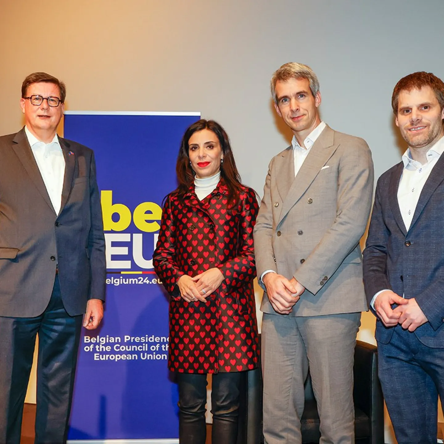 Vier Personen stehen nebeneinander vor einem Banner mit der Aufschrift "Belgian Presidency of the Council of the European Union". Sie tragen formelle Kleidung und blicken in die Kamera.