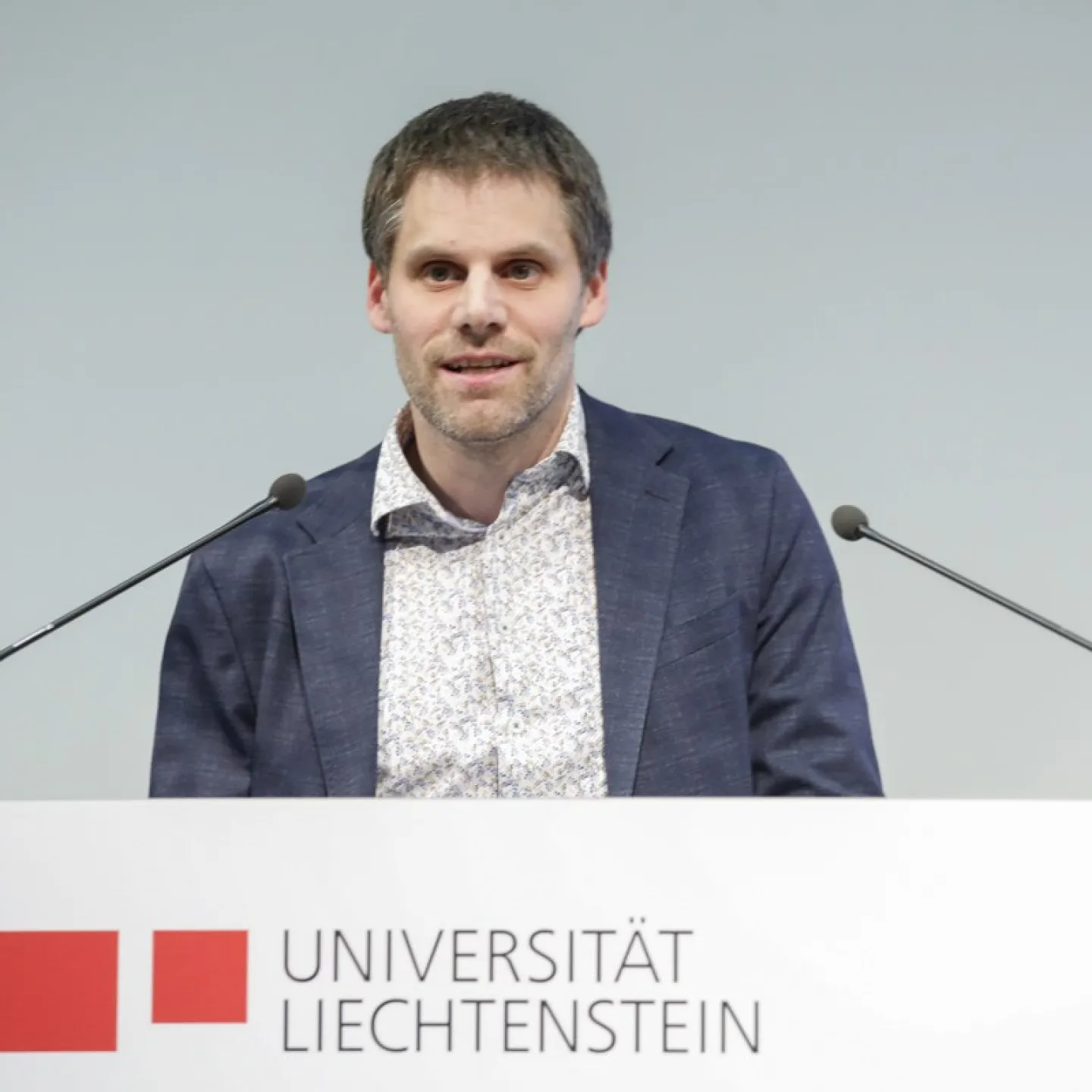 Rektor Christian Frommelt hält eine Rede am Rednerpult vor einem sitzenden Publikum in einem Konferenzraum. Im Hintergrund ist ein Poster sichtbar.