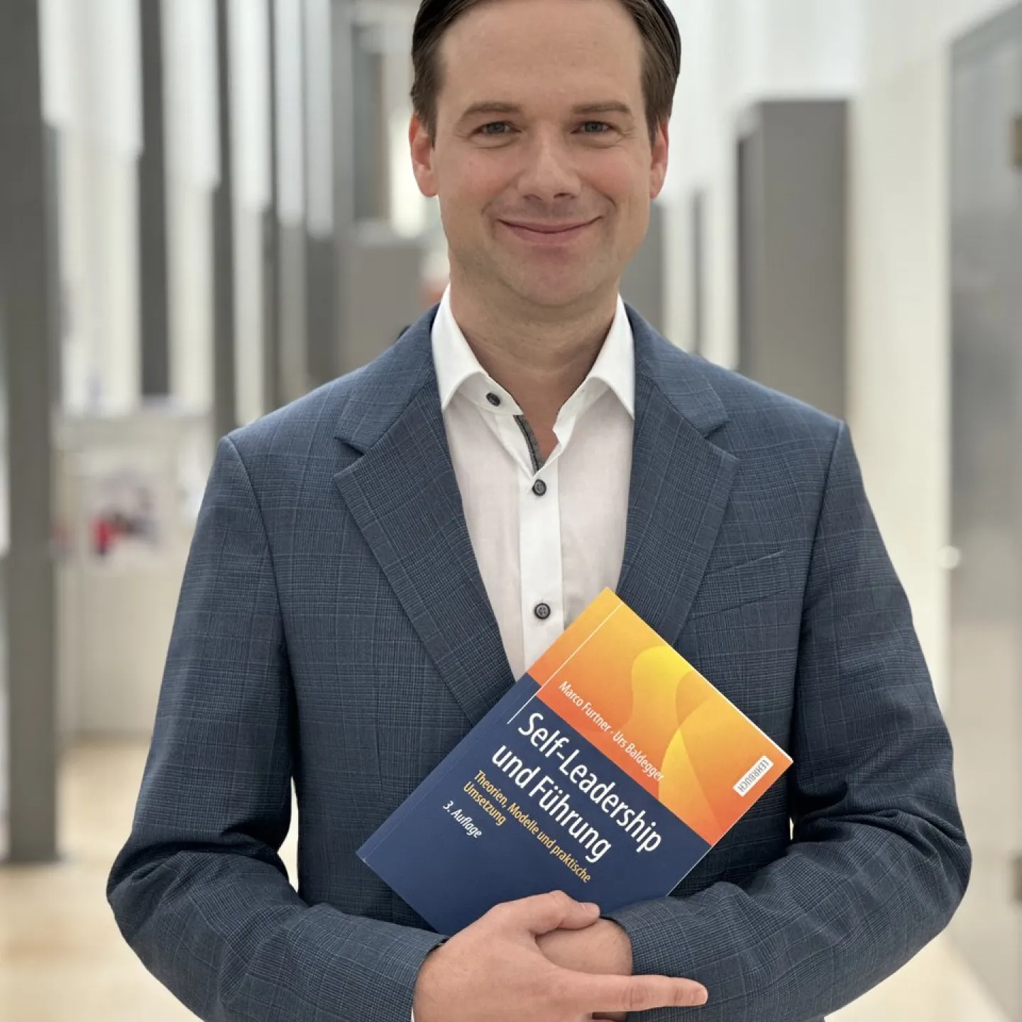 Ein Mann in einem blauen Anzug hält das Buch „Self-Leadership und Führung“ in einem modernen, hellen Flur.