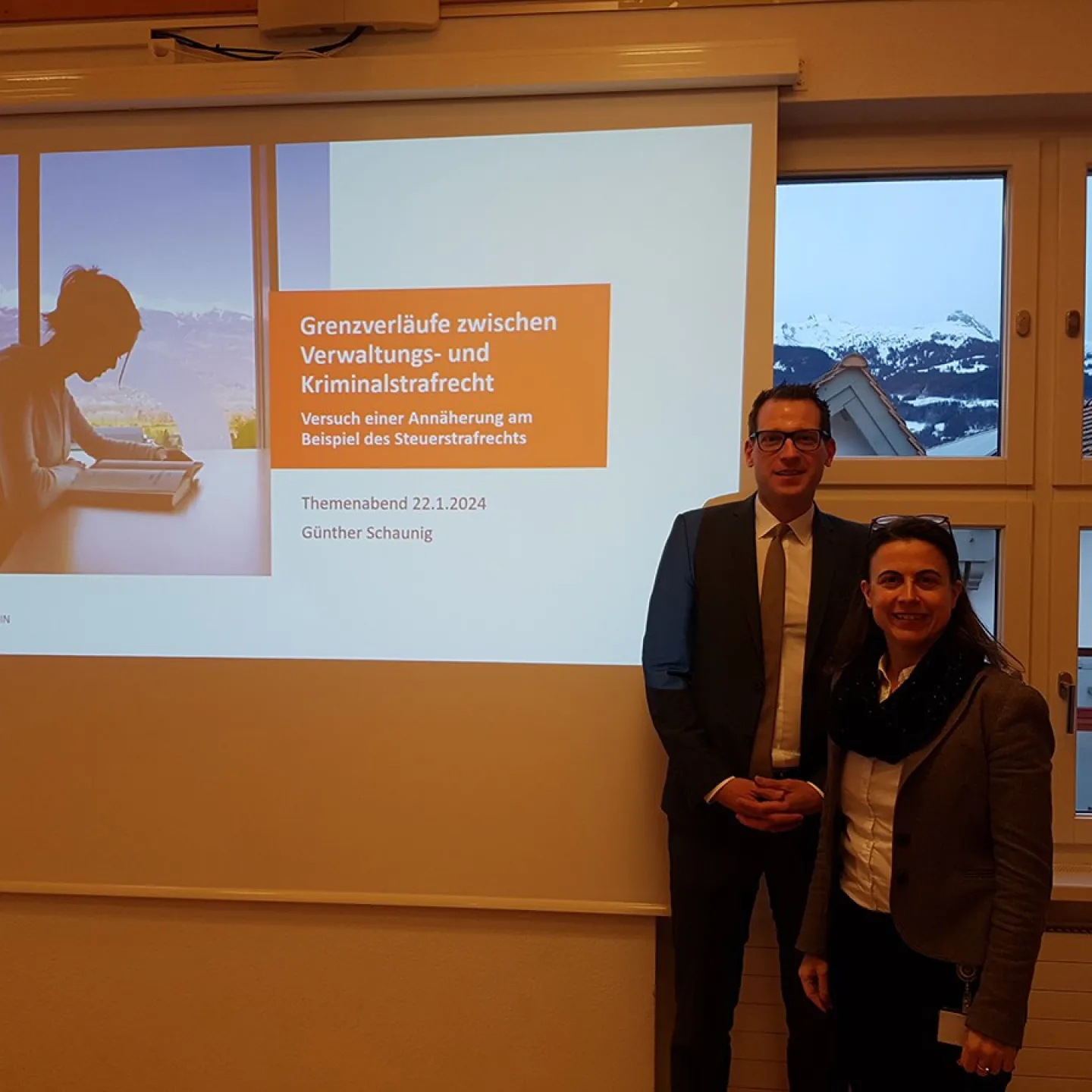 Ein Mann und eine Frau stehen vor einer Präsentationsleinwand mit dem Titel "Grenzverläufe zwischen Verwaltungs- und Kriminalstrafrecht" im Seminarraum der Universität Liechtenstein. Im Hintergrund sind schneebedeckte Berge durch die Fenster sichtbar.