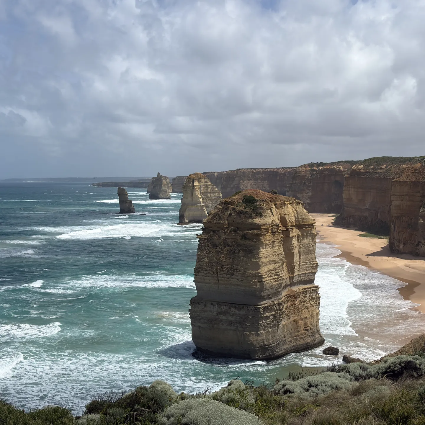 Steile Felsklippen und markante Felsnadeln ragen aus dem Meer entlang einer wilden Küste mit Sandstrand und brechenden Wellen.