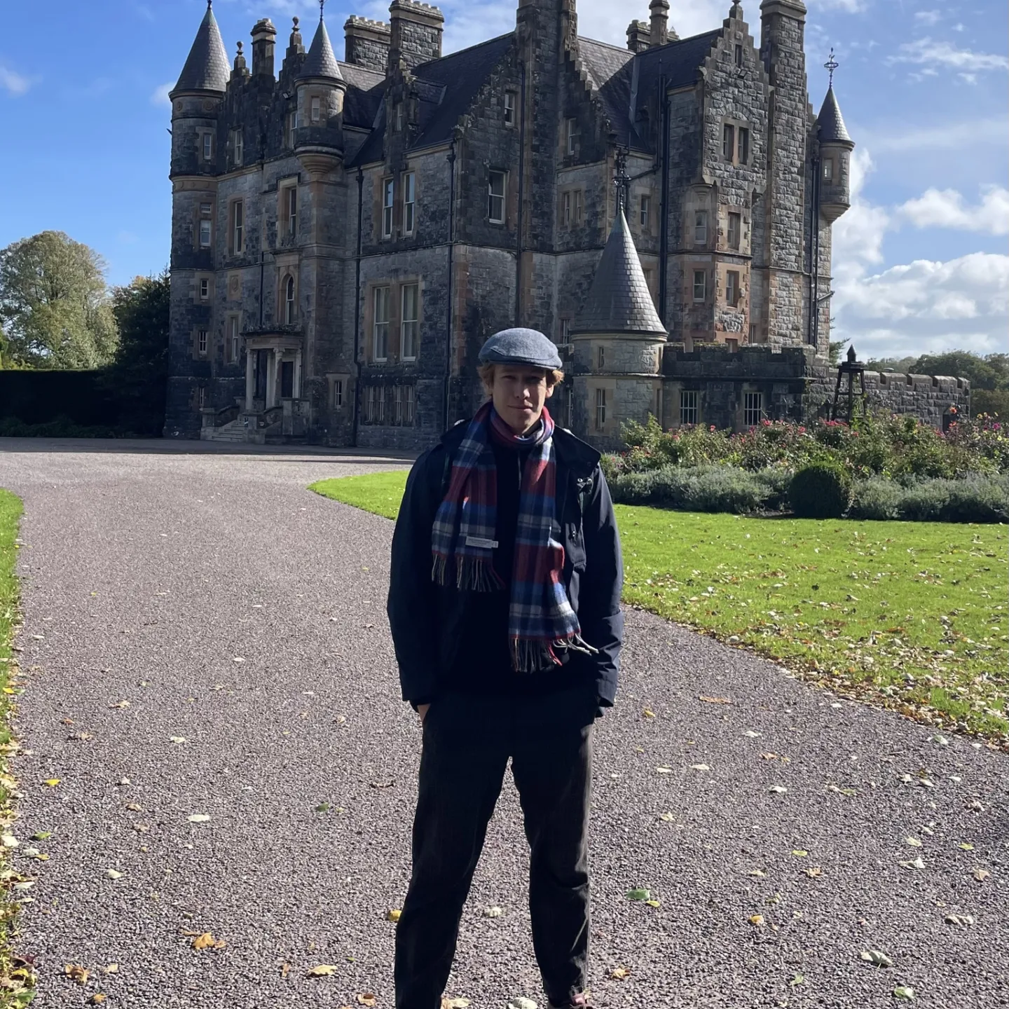 Person mit Mütze und Schal steht auf einer Auffahrt vor einem historischen Schloss aus Stein unter blauem Himmel.