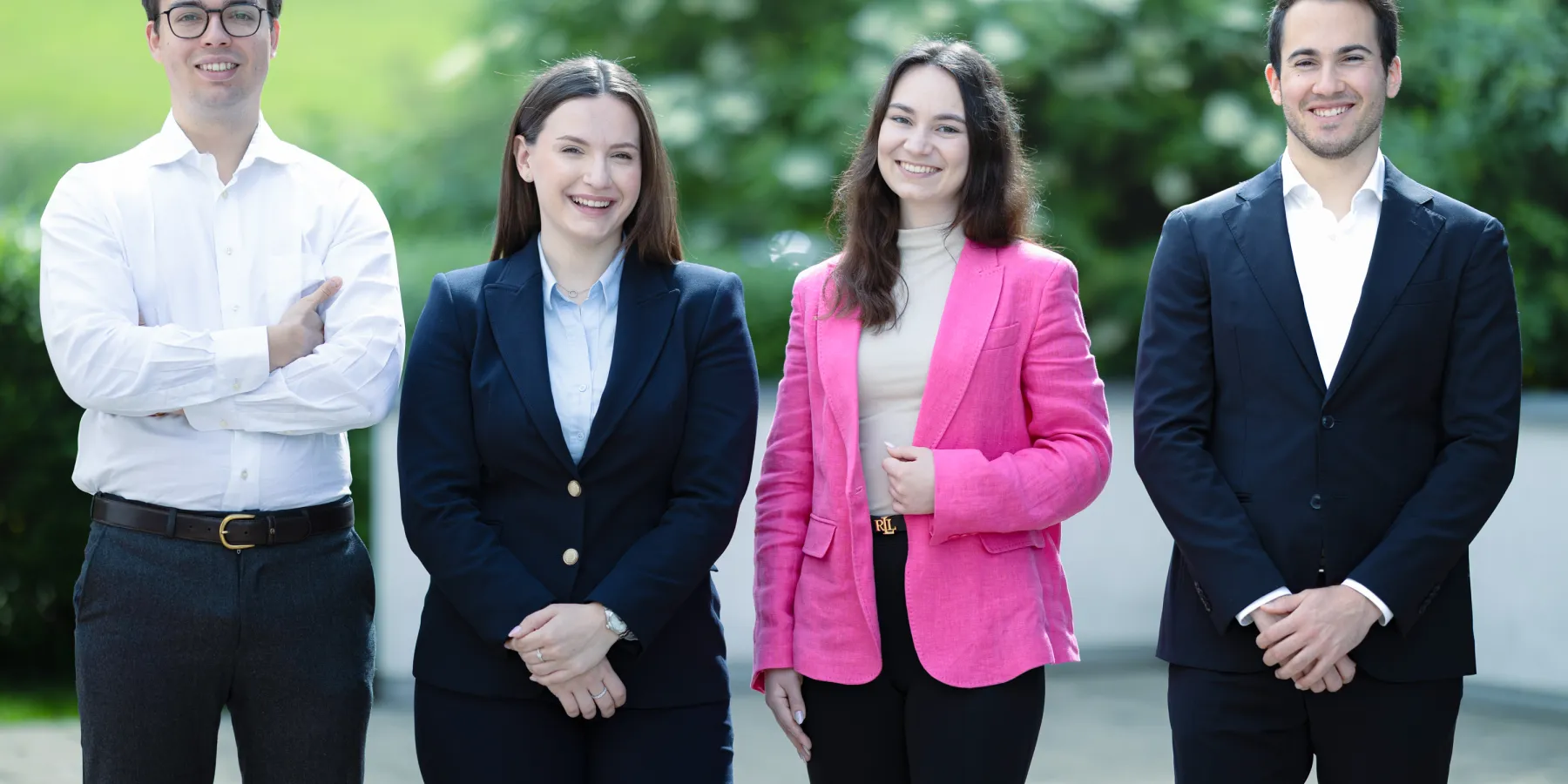 Vier Studierende im Masterstudiengang Innovative Finance der Universität Liechtenstein stehen nebeneinander im Freien, professionell gekleidet und lächelnd in die Kamera. Im Hintergrund ist grüne Natur zu sehen.