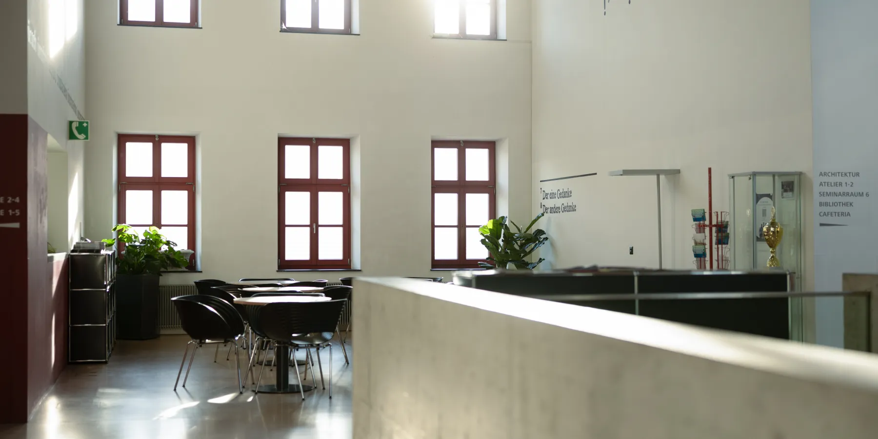 Innenbereich mit hoher Decke und Cafeteria-Atmosphäre, Tageslicht durchflutet Raum mit roten Fenstern und Glasvitrine mit Pokal.