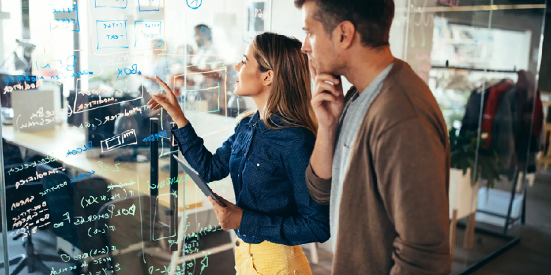 Ein Mann und eine Frau betrachten gemeinsam eine große Glaswand, die mit Notizen und Diagrammen beschrieben ist. Beide wirken nachdenklich und konzentriert, während sie an einer komplexen Aufgabe arbeiten.