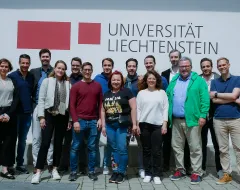 Alumni Gruppe posiert gemeinsam vor dem Logo der Universität Liechtenstein bei sonnigem Wetter auf dem Campus