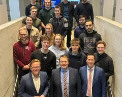 Gruppe von Studierenden und Lehrenden steht auf einer Treppe in einem modernen Gebäude der Universität Liechtenstein, lächelnd in die Kamera. Im Hintergrund sind Bildschirme und eine Steinwand zu sehen.