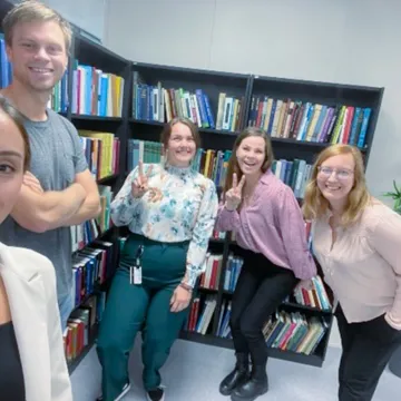 Fünf Mitglieder des International Office der Universität Liechtenstein posieren lachend vor einem Bücherregal in einer Bibliothek.