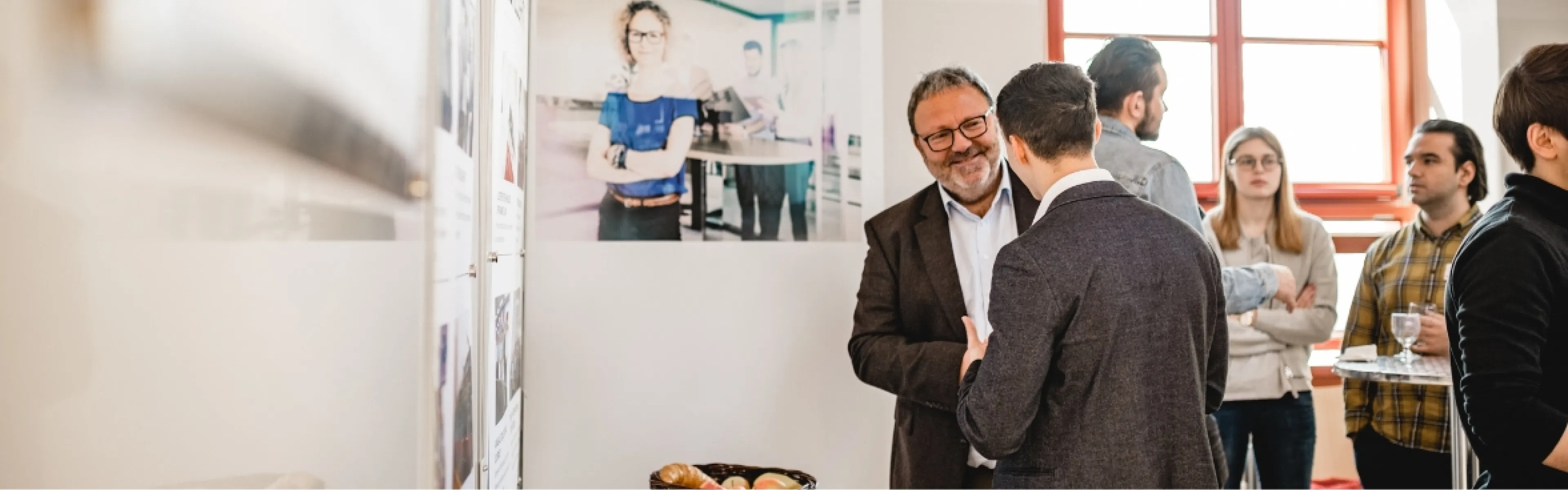 Zwei Männer stehen im Vordergrund und unterhalten sich, während auf der linken Seite Ausstellungsstücke zu sehen sind.