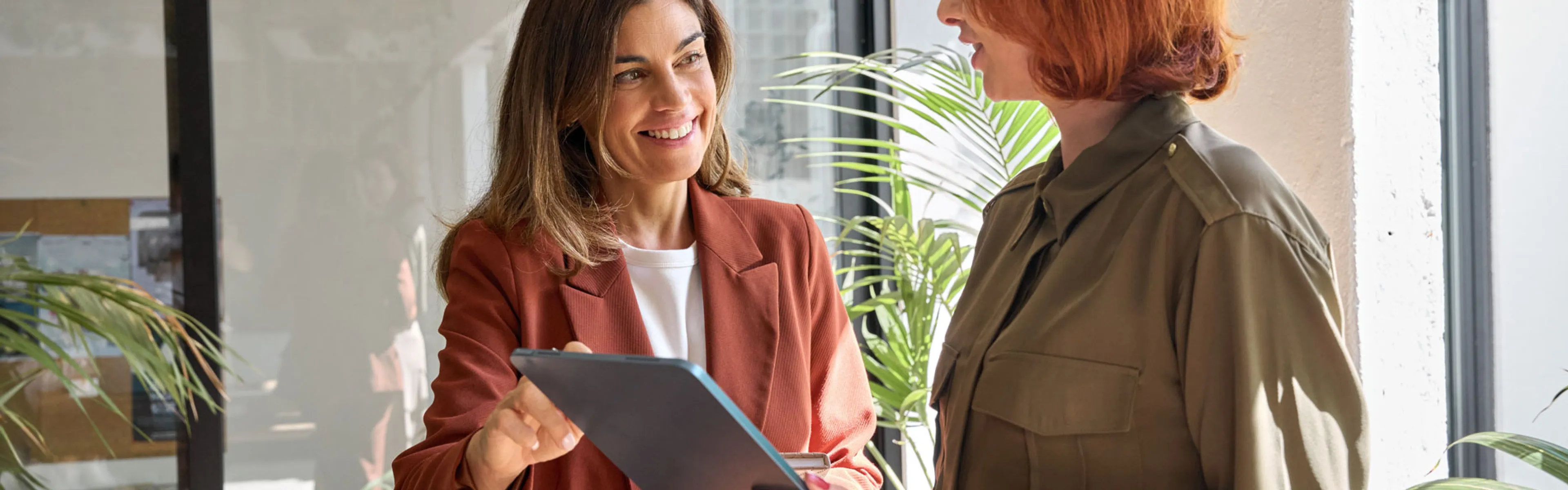 Zwei Frauen stehen in einem modernen Bürogebäude und unterhalten sich. Beide sind formell gekleidet und wirken konzentriert. Im Hintergrund sind große Fenster zu sehen, die viel natürliches Licht in den Raum lassen, sowie Büromöbel und Pflanzen, die eine professionelle Atmosphäre schaffen.