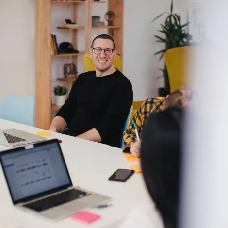 Ein Mann in einem schwarzen Pullover lächelt während eines Treffens in einem Büro. Im Vordergrund sind Laptops und Notizen auf einem Tisch.