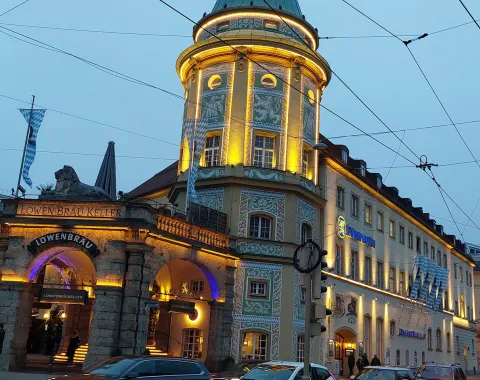 Außenansicht des Löwenbräukellers in München bei Dämmerung, beleuchtet mit warmem Licht, mit vorbeifahrenden Autos und Straßenbahnkabeln im Vordergrund.