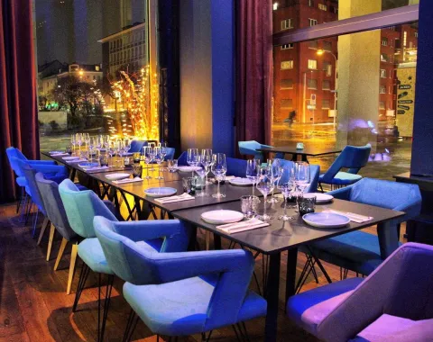 Ein stilvoll gedeckter Tisch in einem modernen Restaurant mit bunten Stühlen und großen Fenstern, die einen nächtlichen Blick auf eine beleuchtete Stadtstraße bieten.